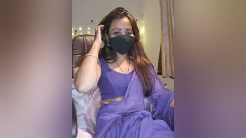 Media: Video of a South Asian woman with long dark hair, wearing a purple saree, black face mask, and headband, sitting on a brown leather chair, in a softly lit, cozy room with fairy lights and curtains.