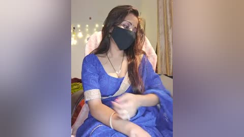 Media: A video of a woman in a blue saree with gold trim, black face mask, and long brown hair, sitting indoors with soft lighting, blurred background.