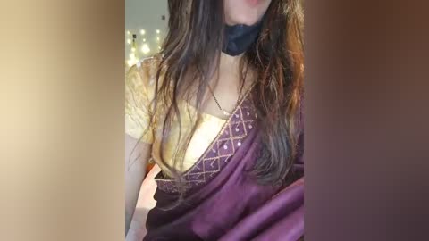 Media: Video of a woman with long, wavy brown hair, wearing a gold blouse with intricate embroidery and a purple sari with golden accents, standing in a dimly lit room with string lights in the background.