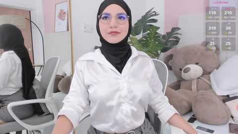 Media: Video of a young woman with light skin, wearing a black hijab, white blouse, and gray skirt, seated at a desk with a laptop, surrounded by a cozy, pastel-themed room with a large teddy bear and a green plant.