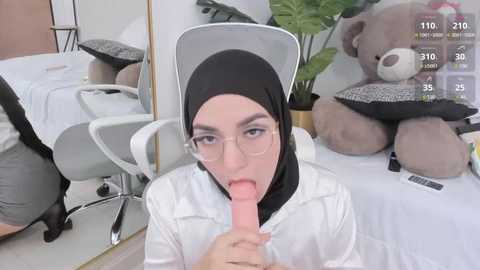 Media: Video of a young woman with glasses, wearing a black hijab and white lab coat, licking a large, erect dildo in a modern office setting with a white chair, green plant, and stuffed animal.