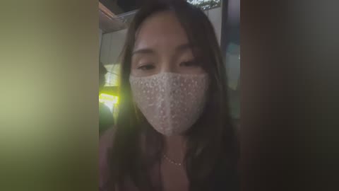 Media: A video of an Asian woman with long brown hair wearing a white face mask, sitting in a dimly lit room, with a blurred background of a vehicle's interior.