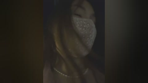 Media: A dimly lit video of an Asian woman with long black hair, wearing a glittery mask, a simple necklace, and a dark top, her eyes closed, giving a mysterious, ethereal vibe.