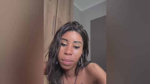 Video of a young Black woman with wet, dark brown hair and full lips, wearing a necklace, standing indoors with beige walls and a wooden cabinet in the background.