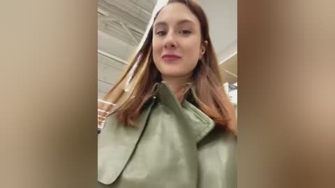 Media: A video of a young woman with light skin and shoulder-length brown hair, wearing a green leather jacket, standing in a modern, indoor setting with beige walls and exposed ceiling beams.