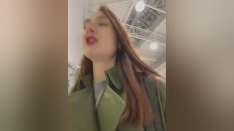 Media: Video of a young woman with straight red hair, wearing a green jacket, red lipstick, and standing in a modern, industrial-style room with white ceiling beams and exposed pipes.