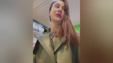 Media: Video of a young woman with fair skin, long brown hair, and red lipstick, wearing a khaki trench coat, in a brightly lit room with white ceiling panels and green walls.