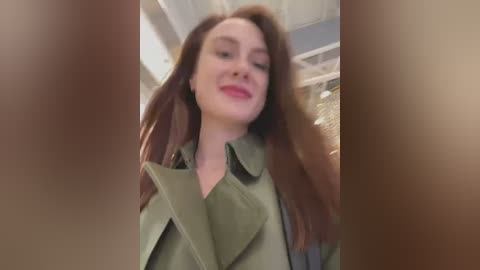 Media: Video of a smiling Caucasian woman with long brown hair, wearing a green trench coat, taken from a low angle, partially obscured by brownish blur.