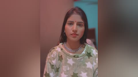 Media: A video of a young Indian woman with medium brown skin, long dark hair, and a calm expression. She wears a green and white star-patterned sweater and a silver necklace. The background is blurred, with warm lighting.