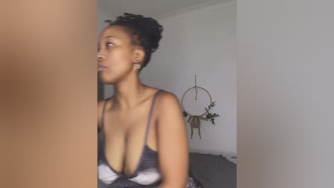 Media: Video of a dark-skinned woman with curly hair, wearing a low-cut black and white tie-dye top, standing in a minimalist bedroom with a wall hanging and a bed.