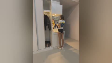 Media: A video of a young woman with a fit physique, wearing a white crop top and black shorts, standing in front of a white wardrobe, looking inside.