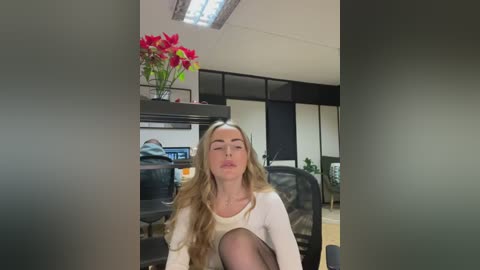 Media: Video of a blonde woman with long, wavy hair, wearing a white top, sitting in an office chair, leaning back with eyes closed, surrounded by office furniture and a floral arrangement on a shelf.