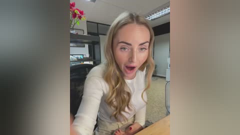 Media: A video of a blonde woman in a white top, surprised expression, sitting at a desk in an office with a vase of red flowers and a black cabinet in the background.