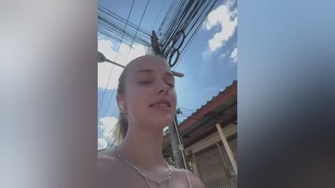 Media: Video of a smiling, fair-skinned woman with blonde hair, wearing a light top, standing in front of an urban scene with power lines and a red-roofed building.