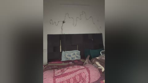 Media: Video of a messy bedroom with a bed covered in pink and red patterned bedding, a green pillow, and a white blanket. The ceiling shows black handprints and a broken light fixture.