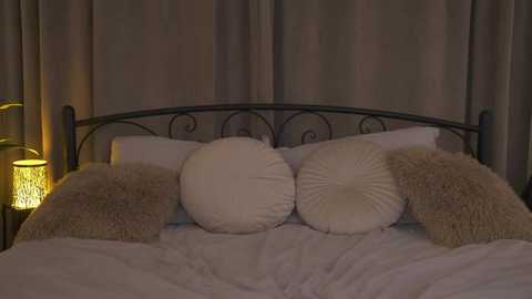 Media: Video of a dimly lit bedroom featuring a black wrought iron bed with two large white cushions, beige throw pillows, and a warm, yellow-lit lamp on the left side.