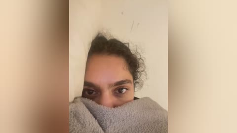 Media: Video of a person with curly dark hair, partially hidden behind a grey towel, peering through a gap in a beige wall.