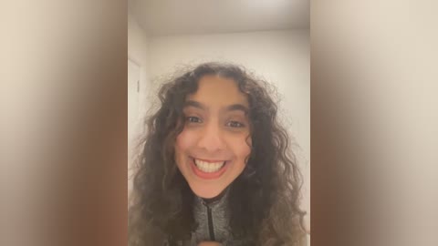 Media: Video of a smiling woman with curly dark hair and light brown skin, wearing a gray top, standing in a narrow hallway with white walls and brown flooring.