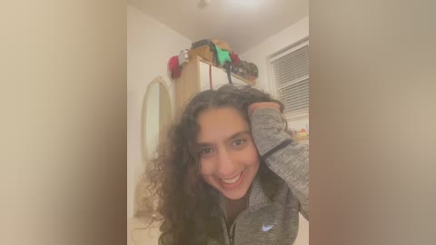 Media: A video of a young woman with curly hair, wearing a grey sweatshirt, smiling and adjusting her hair in a brightly lit room with beige walls, a small window, and a wooden dresser.