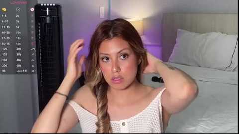 Media: Video of a young Latina woman with long, wavy hair, wearing a white lace top, sitting on a bed in a dimly lit bedroom.