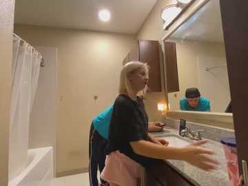 Media: A video of a blonde woman in a black top, pink apron, and turquoise apron, cleaning a bathroom counter. The bathroom has beige walls, a white bathtub, and a dark wooden vanity mirror.