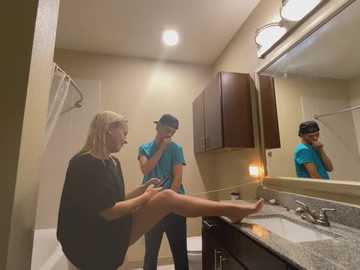 Media: Video of a blonde woman in a black top sitting on a bathroom counter, leaning back against a man in a blue shirt, both laughing, with a mirror reflecting a third man in the background.