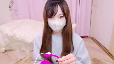 Media: Video of an Asian woman with long brown hair and a light blue mask, holding a pink vibrator, in a pastel-toned, softly lit bedroom.