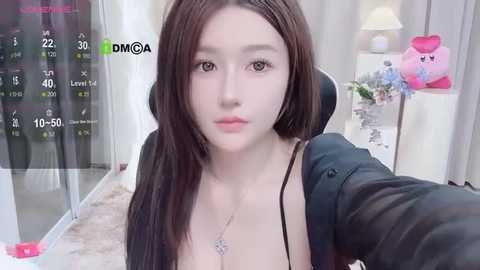 Media: A video of an East Asian woman with long, straight black hair, fair skin, and medium-sized breasts, wearing a black top and a silver necklace, sitting in a modern living room with a large window, a pink lamp, and a white lampshade.