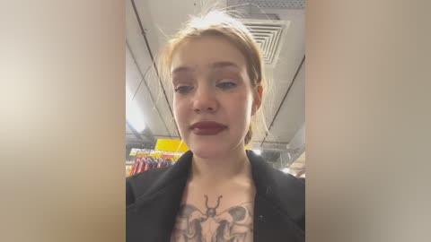 Media: Video of a young Caucasian woman with fair skin, blonde hair, and red lipstick, wearing a black jacket and revealing a detailed tattoo of deer antlers on her chest. The background shows a supermarket aisle with fluorescent lights.