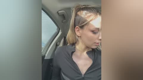 Media: Video of a young woman with light skin and light blonde hair, pulled back in a ponytail, wearing a black zip-up top, sitting in a car.