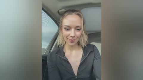 Media: Video of a smiling, light-skinned woman with shoulder-length blonde hair, wearing a black zip-up hoodie, sitting in a car with gray interior.