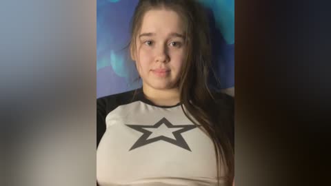 Video of a young woman with fair skin and long brown hair, wearing a black-and-white Star Wars T-shirt. She has a neutral expression. Background is a gradient of blue and purple hues.