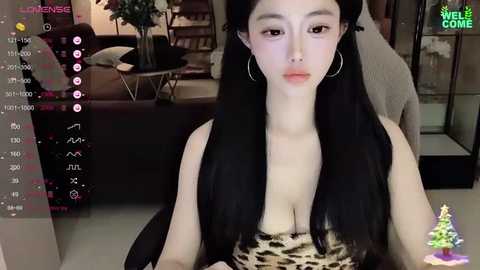 Media: Video of an East Asian woman with long black hair, fair skin, and large breasts, wearing a leopard-print top, sitting indoors. Background includes a couch, potted plants, and a green tree ornament.