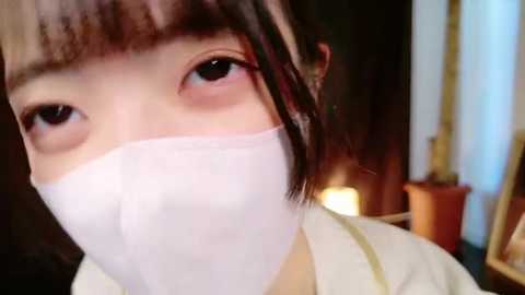 Media: Video of a young East Asian woman with fair skin, wearing a white surgical mask, and brown hair with bangs, indoors with warm lighting and wooden decor.