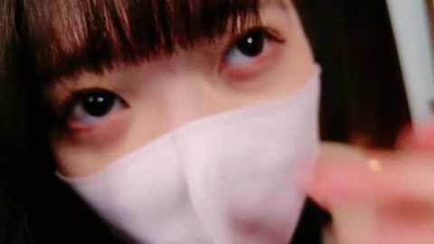 Media: Video of an Asian woman with straight black hair, wearing a white surgical mask, looking up with wide eyes, blurred background.