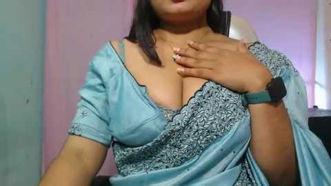 Media: Video of a South Asian woman with medium brown skin, wearing a light blue off-shoulder blouse with intricate lace and a matching skirt. She has a large bust and is wearing a smartwatch on her left wrist. The background features a pink wall and a white lamp.