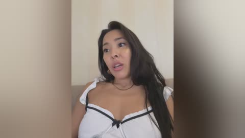 Media: Video of a young, dark-haired Asian woman in a white, low-cut maid outfit, with a surprised expression, against a plain beige background.