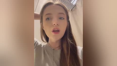 Media: Video of a young, fair-skinned woman with long, straight brown hair, wearing a grey top, with a surprised expression. Background features a beige wall and a window with blinds.