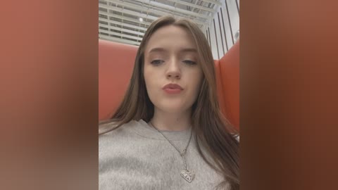 Media: Video of a young woman with long brown hair, light skin, and pink lipstick, wearing a gray sweatshirt and silver necklace, seated indoors with orange walls and white ceiling.