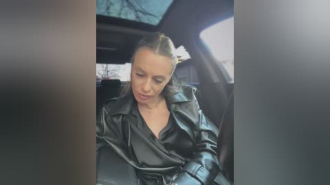 Media: A video shows a blonde woman with a ponytail, dressed in a black leather jacket, seated in a car. The interior is dimly lit, with blurred windows and a tree branch visible outside.