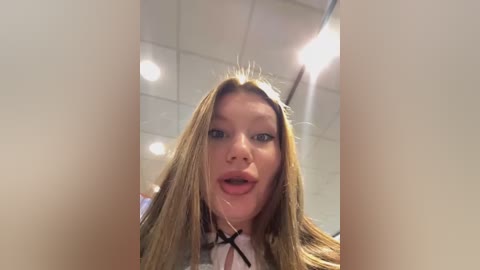 Media: Video of a young woman with long, blonde hair and fair skin, wearing a white collared shirt. She is indoors, possibly in an office with fluorescent lights. Her expression is open-mouthed and surprised.