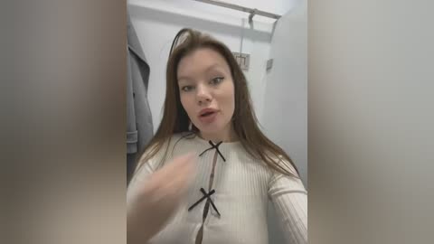 Media: Video of a young Caucasian woman with long brown hair, wearing a white ribbed sweater with black bow accents, standing in a laundry room with white walls and hanging clothes.