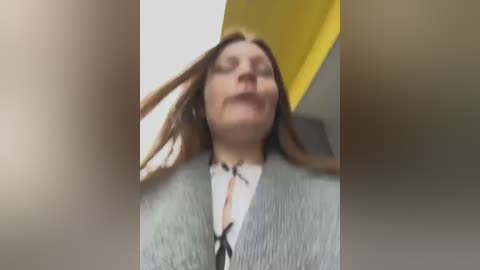 Media: Video of a woman with long brown hair, wearing a grey cardigan over a white blouse, standing in a yellow hallway, looking upwards with a slight smile.