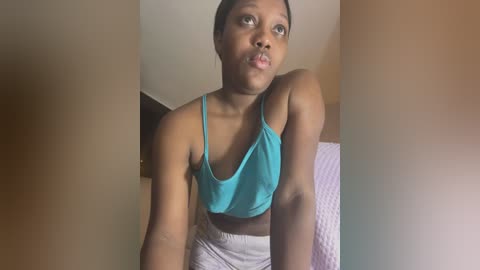 Media: Video of a young Black woman with short hair, wearing a turquoise tank top and gray pants, gazing upward with a contemplative expression. Background features a beige wall and a pink quilted blanket.