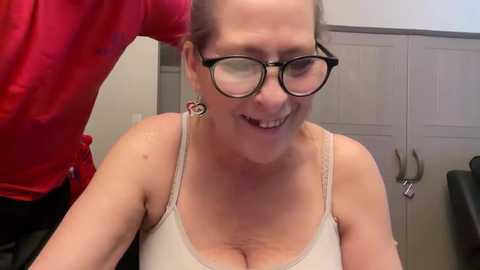 Media: Video of a cheerful, middle-aged woman with glasses, wearing a beige tank top, seated in a room with grey cabinetry and a red cloth visible in the background.