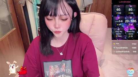 Media: A video of a young Asian woman with straight black hair and fair skin, wearing a maroon t-shirt featuring a cartoon, sitting on a beige couch. The background shows a wooden door and a TV screen displaying a live stream.