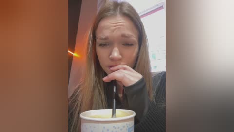 A video of a young woman with long brown hair, wearing a black sweater, drinking from a white coffee cup with a black straw, looking slightly worried, set in a dimly lit room with red and white walls.