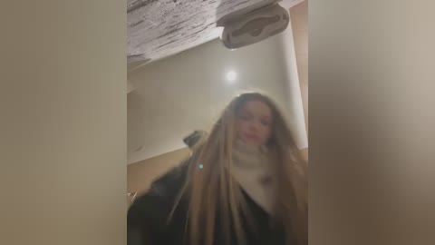 A blurred video shows a young woman with long, blonde dreadlocks, wearing a black coat, standing in a dimly lit room with a wooden ceiling and beige walls.