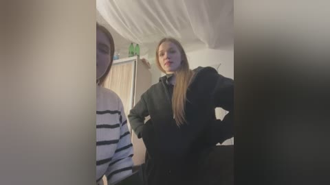 Media: Video of two young women, one with long straight hair, wearing a black hoodie, standing in a bedroom with white drapes, a green bottle, and wooden furniture.