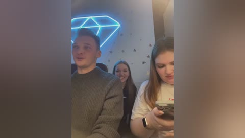 Media: Video of three young people smiling in a dimly lit room with a neon blue diamond-shaped sign and star-patterned wallpaper. One person is wearing a gray sweater, another a white shirt, and the third has a black phone.